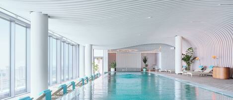A modern indoor swimming pool with a curved ceiling, large windows, and lounge chairs along the poolside.