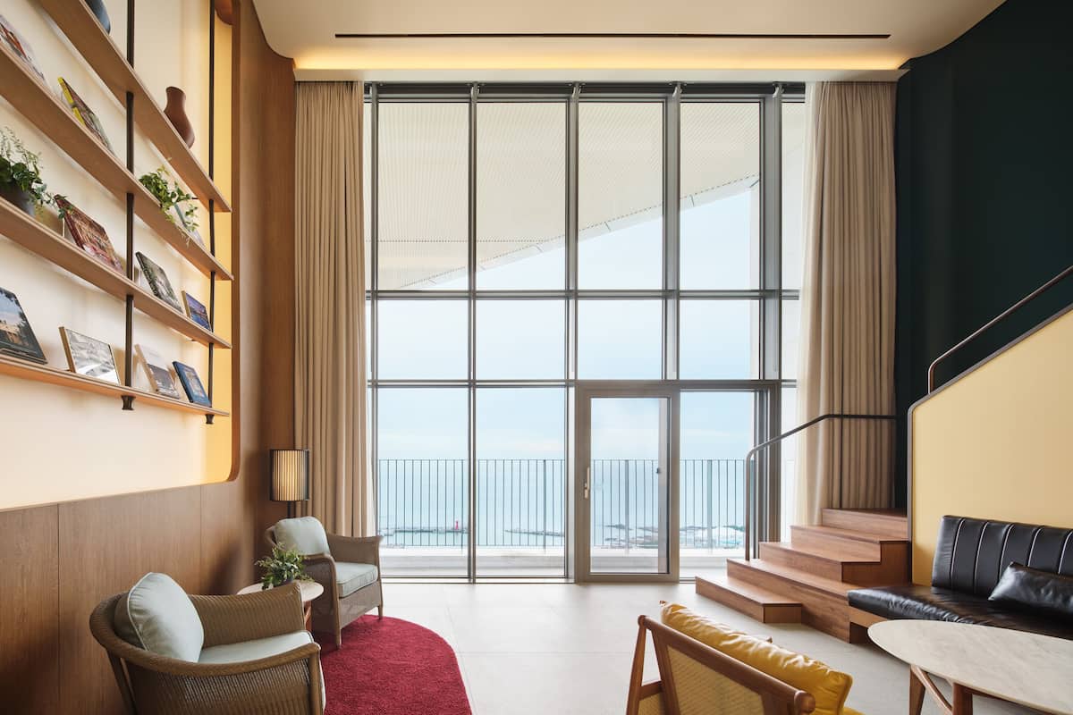A spacious living area with a large window, a bookshelf, a red rug, and a staircase leading to an outdoor balcony.