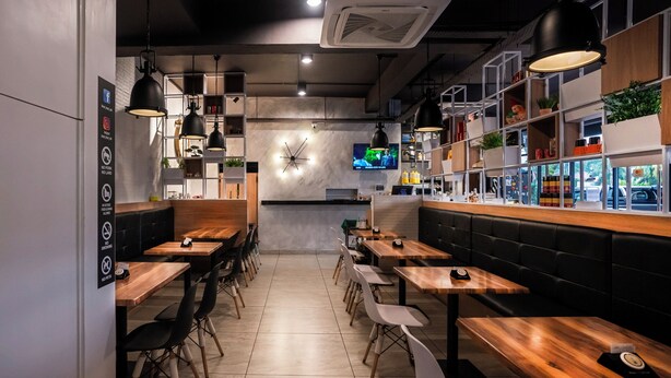 A modern restaurant interior with wooden tables, black and white chairs, pendant lighting, and a TV mounted on the wall. - Hope Hotel (Masai)