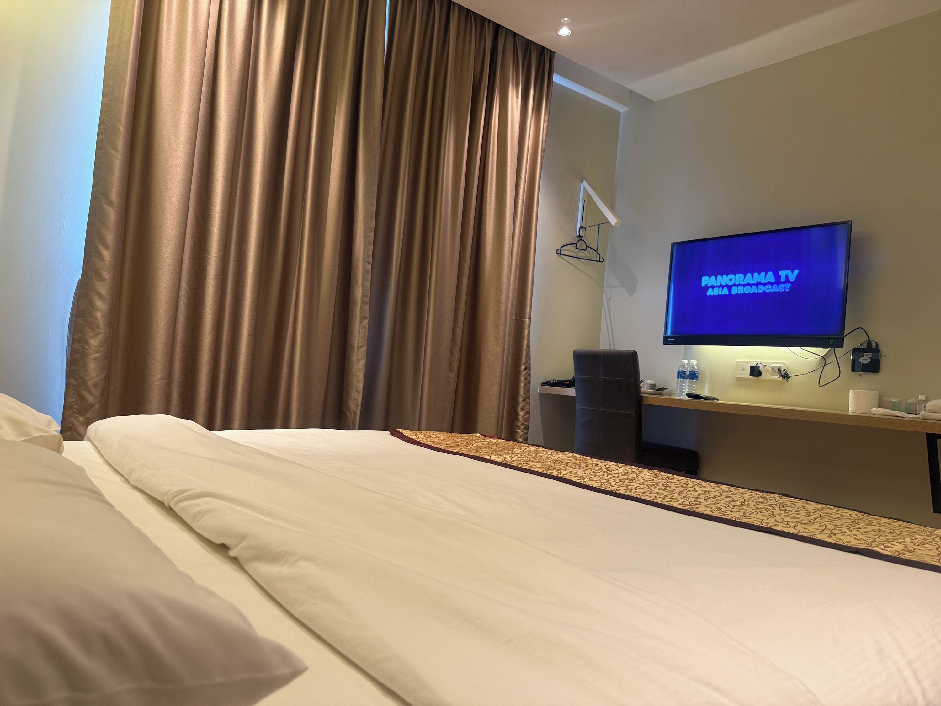 A hotel room with a bed, a desk with a television displaying 'MANORAMA TV ASIA BROADCAST', and a window with curtains.