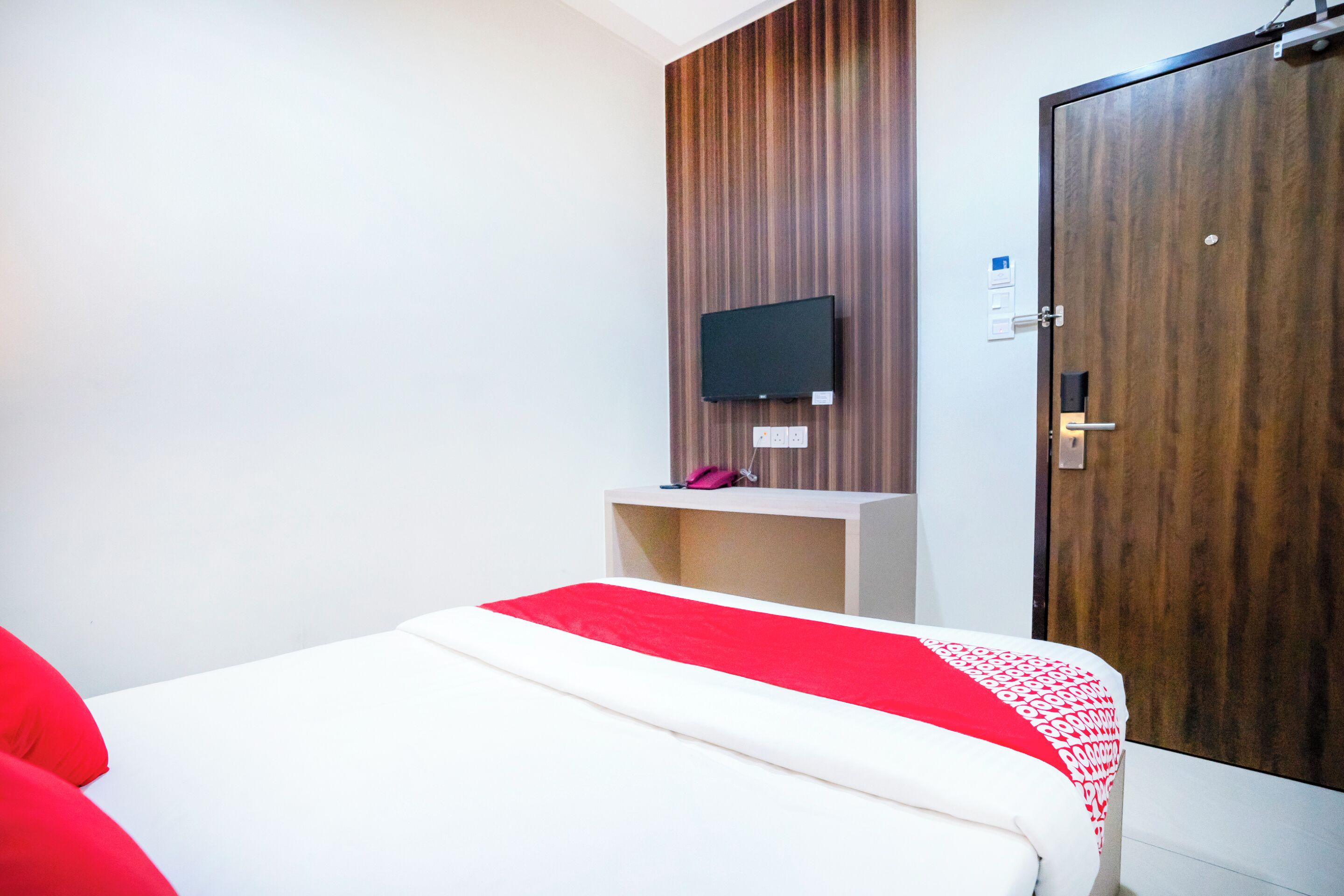 A hotel room with a bed, a television mounted on a wooden panel wall, a door, and a small shelf with a pink item.