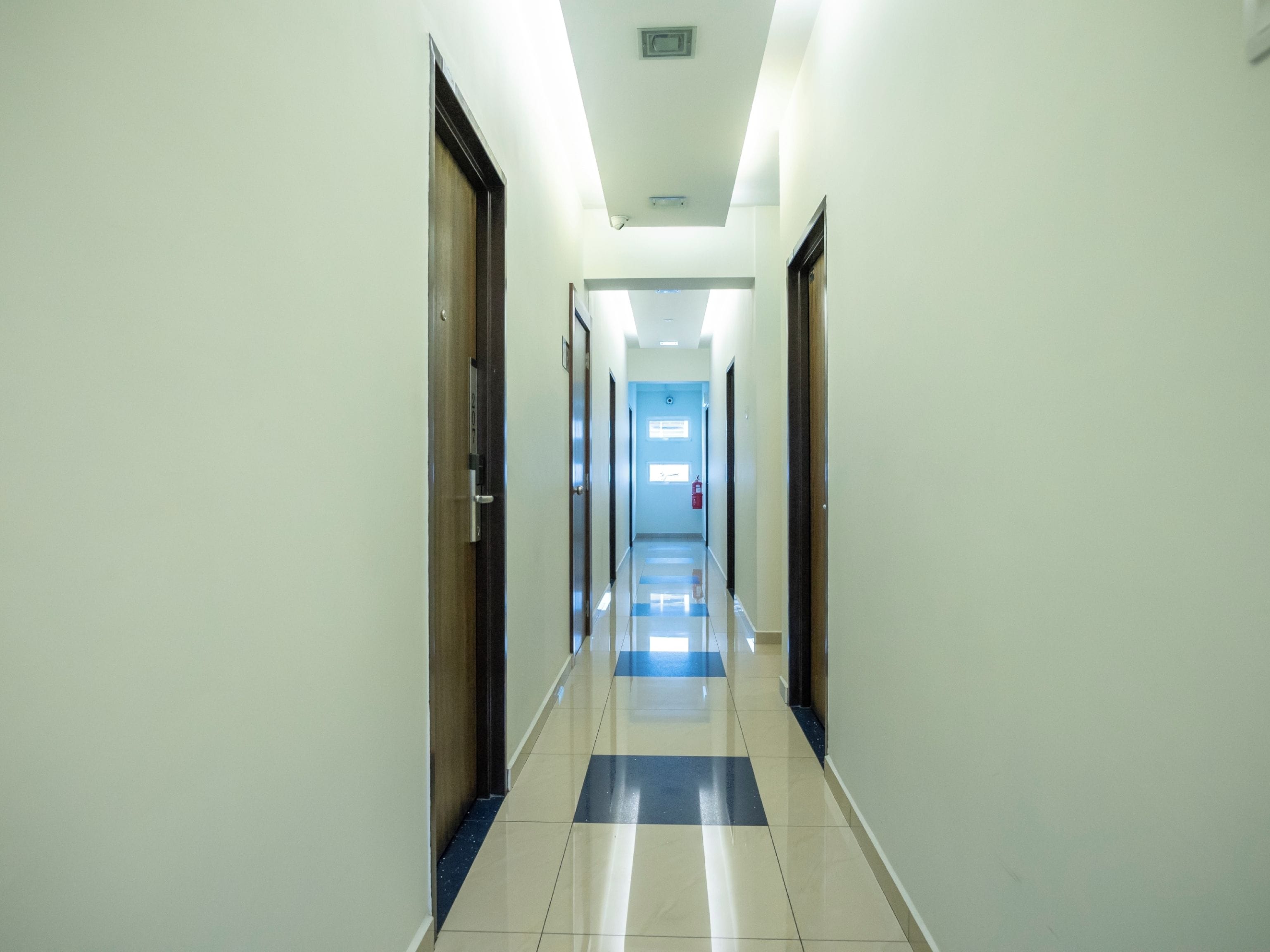 A long, narrow hallway with doors on both sides, a tiled floor with a blue and white pattern, and a ceiling with recessed lighting.