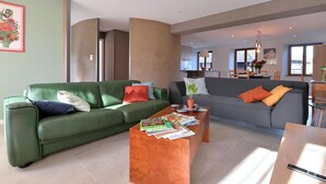 A modern living room with a green sofa, a grey sofa, and a wooden coffee table with magazines. There is a kitchen in the background with a dining table and chairs. - Charming renovated apartment for 4 with terrace in Hunawihr (Hunawihr)