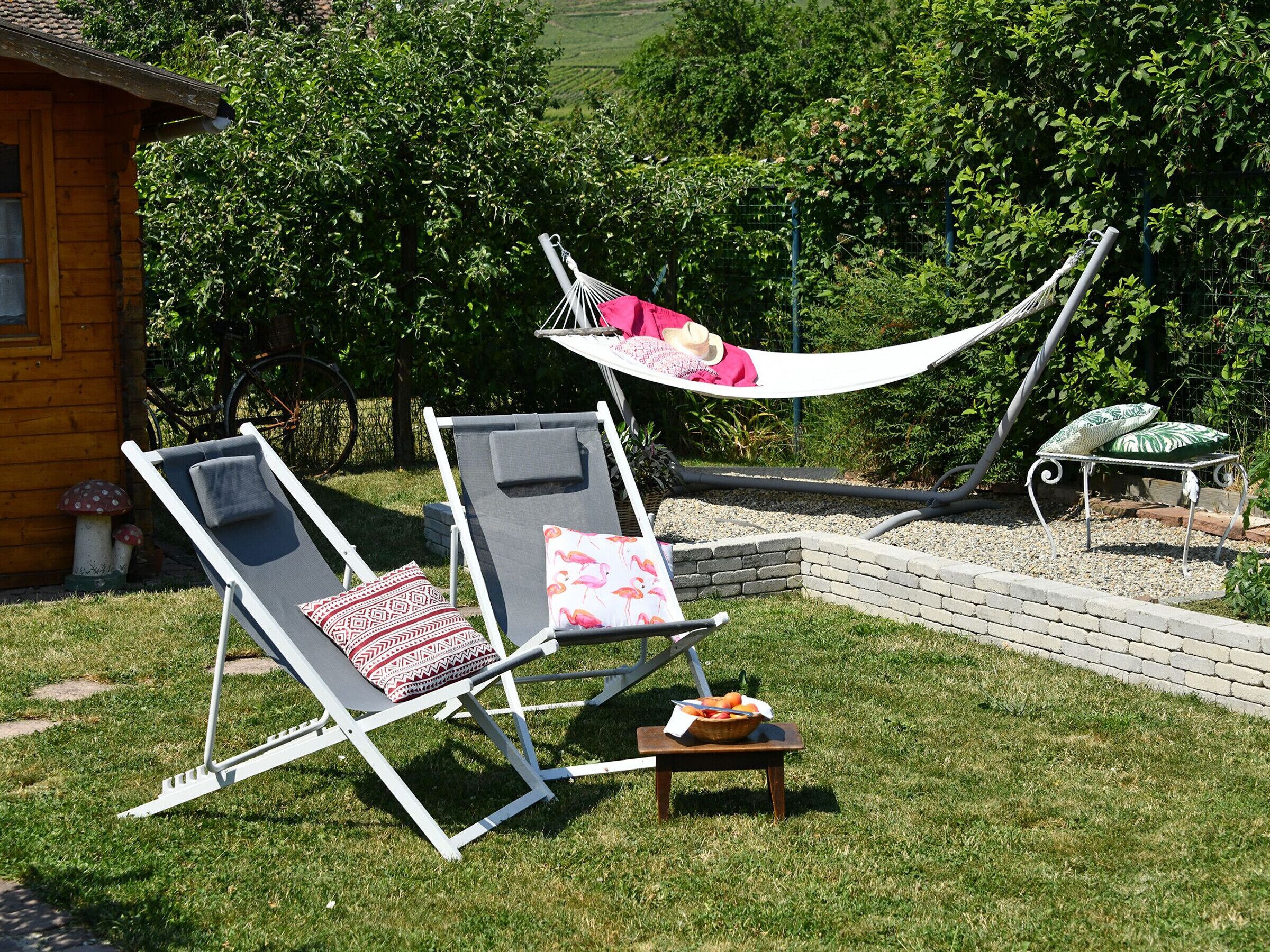 Eine Hängematze zwischen zwei Liegestühlen in einem Garten mit einem Tisch und einer Korb voller Früchte.