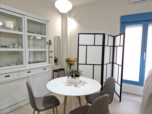 Un comedor con una mesa redonda blanca, cuatro sillas grises, un armario blanco con puertas de vidrio, una separador negro y una ventana con detalles azules.