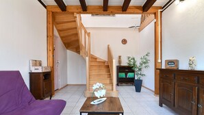 A living room with a purple sofa, wooden staircase, a small table with a white vase, and a cabinet with decorative items. - Beautiful Country Home with Terrace, Close to Amenities & Highways, Sleeps 5 + Baby (Baldersheim)