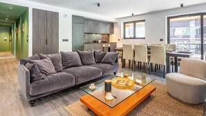 A modern living room with a grey sofa, a glass coffee table, and a dining area with chairs and a table. - Luxurious Apartment with WiFi & Garage - Ski-In/Ski-Out, New Residence (Val-d'Isère)
