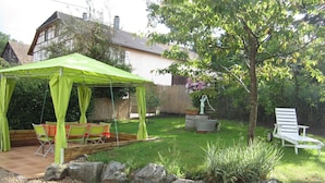 A covered outdoor seating area with a table and chairs, a garden with a tree and a bench, and a house in the background. - Charming Alsatian Farmhouse: 2 Terraces, BBQ, Accessible, Near Vosges – Sleeps 6 (Bretten)