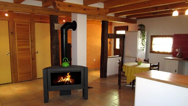 A cozy room with a wood-burning stove, wooden beams on the ceiling, a dining area with a table and chairs, and a kitchen area with a refrigerator and sink. - Charming Alsatian Farmhouse: 2 Terraces, BBQ, Accessible, Near Vosges – Sleeps 6 (Bretten)