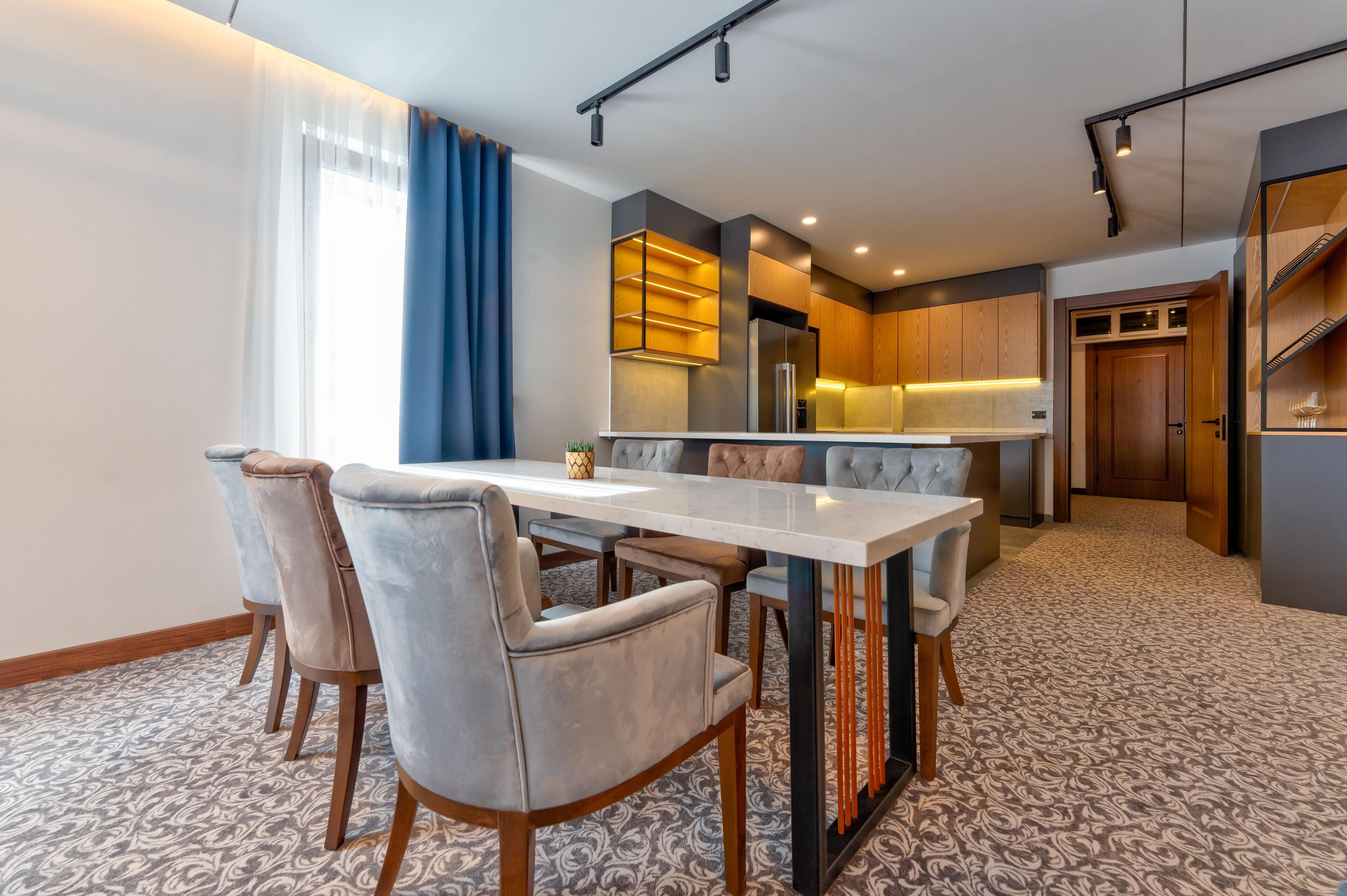 A modern hotel room with a dining area featuring a marble table and cushioned chairs, a kitchen with wooden cabinets and a built-in oven, and a patterned carpet.