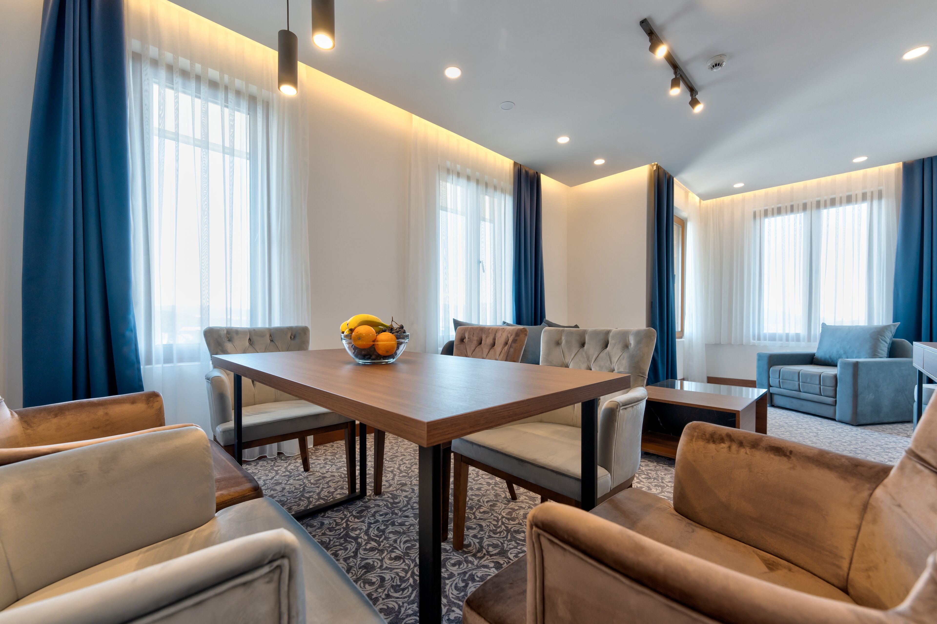 Una habitación de hotel moderna con zona de comedor, sofá y un amplio ventanal con cortinas.