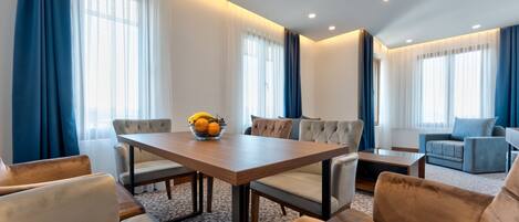 Una habitación de hotel moderna con zona de comedor, sofá y un amplio ventanal con cortinas.