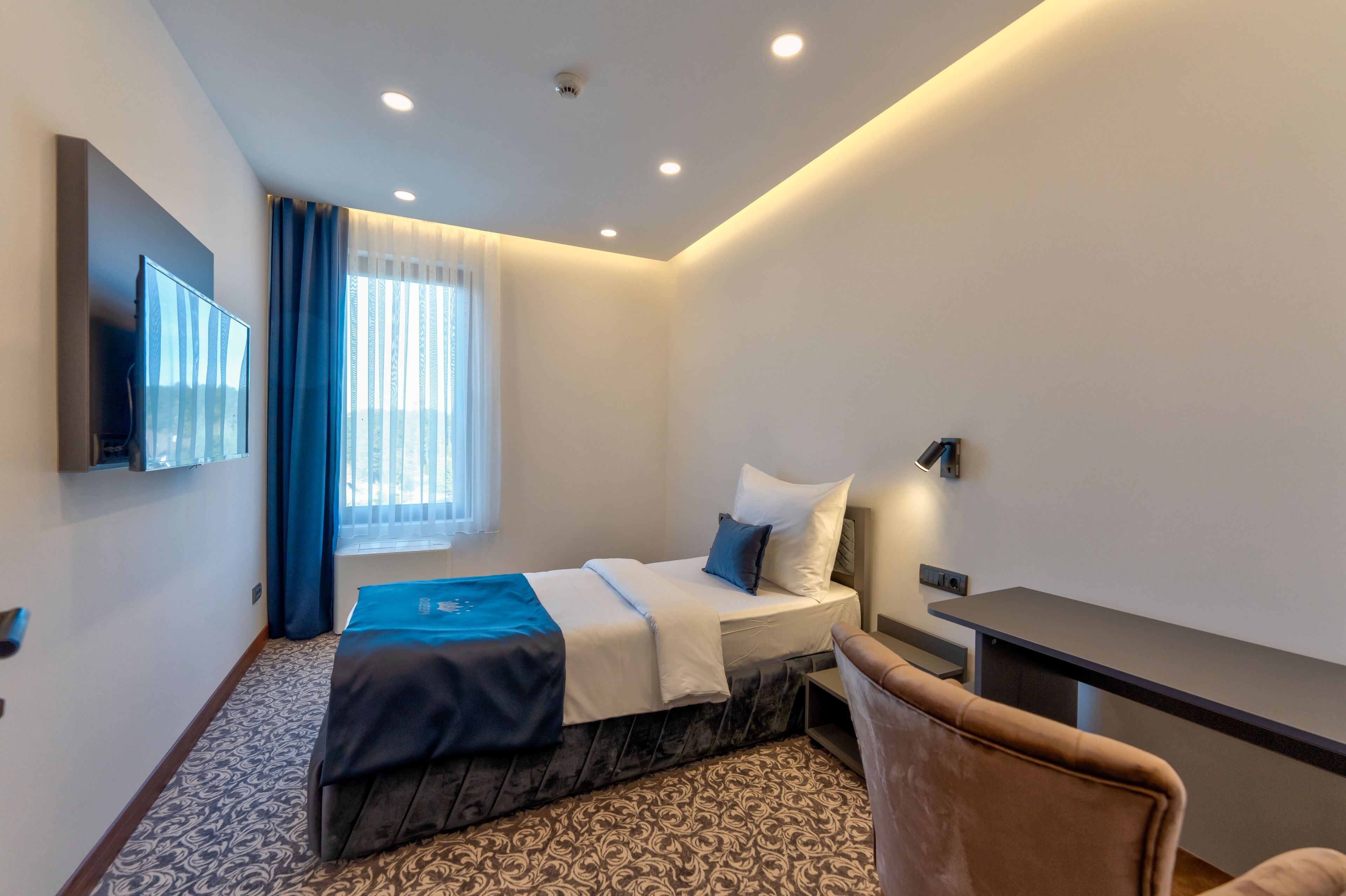 A modern hotel room with a neatly made bed, a flat-screen TV mounted on the wall, a desk with a chair, and a window with curtains.