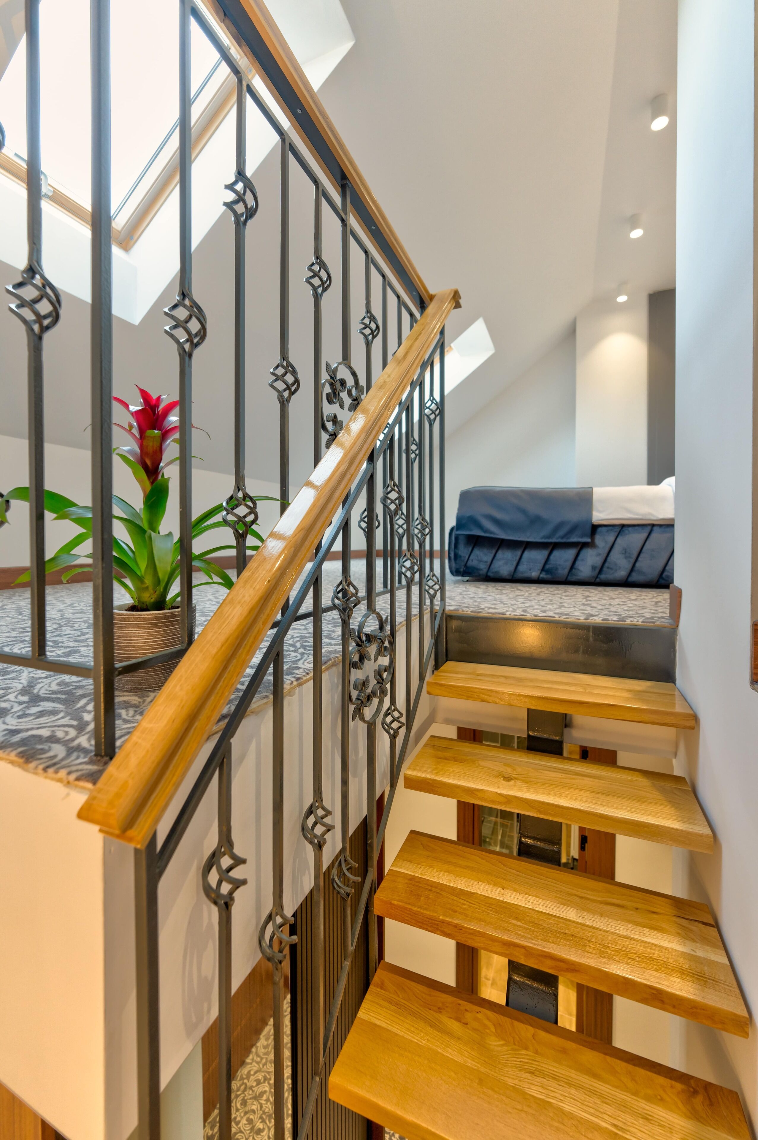 Un escalier moderne avec des marches en bois et des rampes en métal, menant à une chambre à coucher équipée d’un lit et d’une plante en pot.