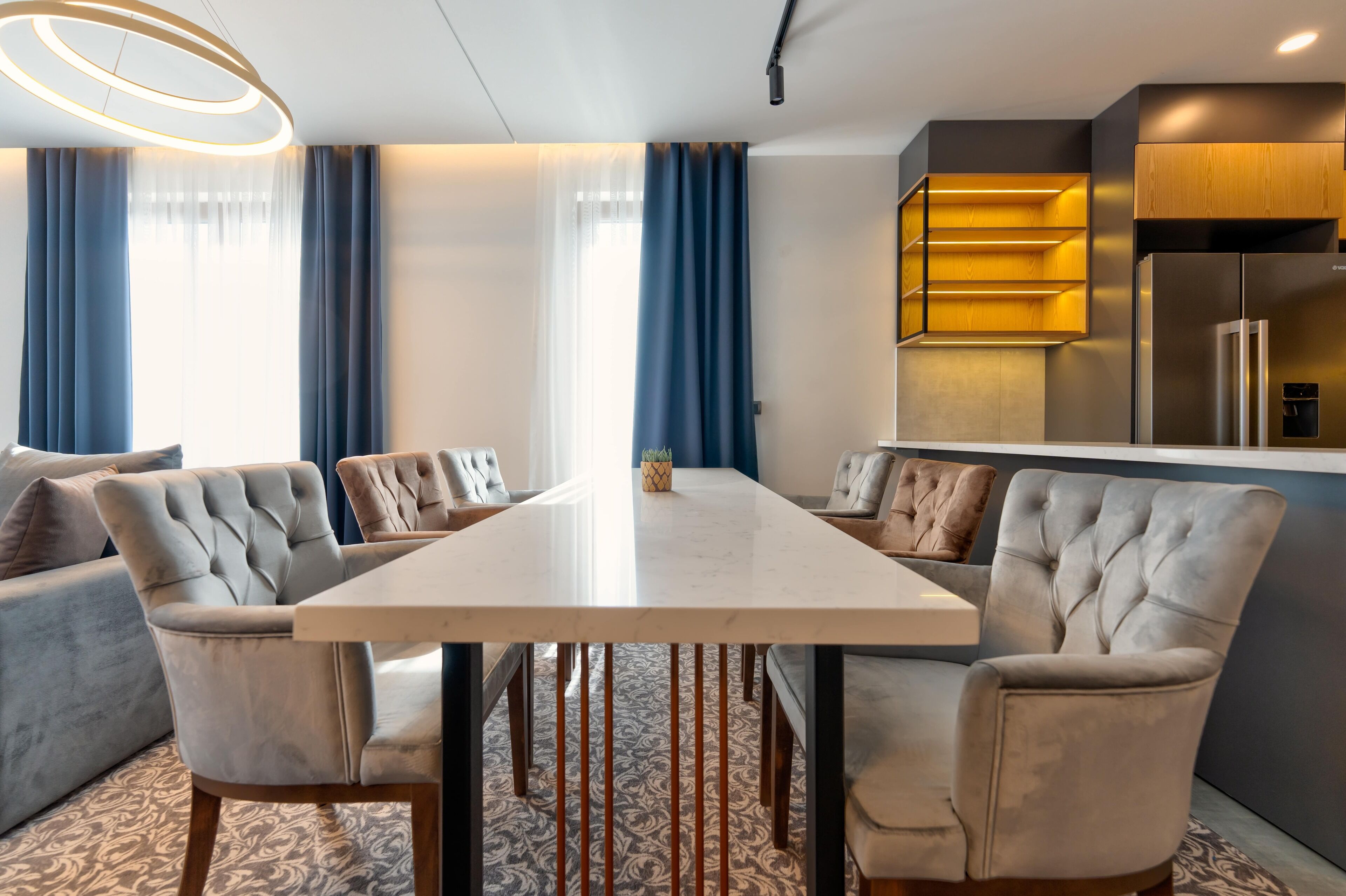 A modern dining area with a large table, upholstered chairs, and a kitchen area with a refrigerator and wooden cabinets.