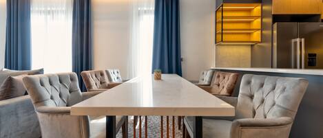 Un comedor moderno con una mesa grande, sillas tapizadas y una zona de cocina con refrigerador y armarios de madera.
