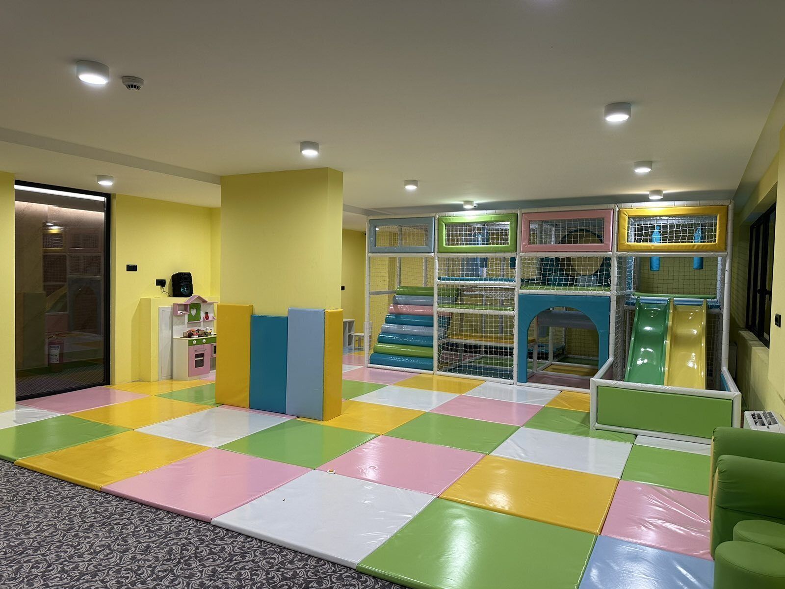 Un área de juegos infantiles con piso acolchado de colores, estructuras de juego y una zona de descanso.