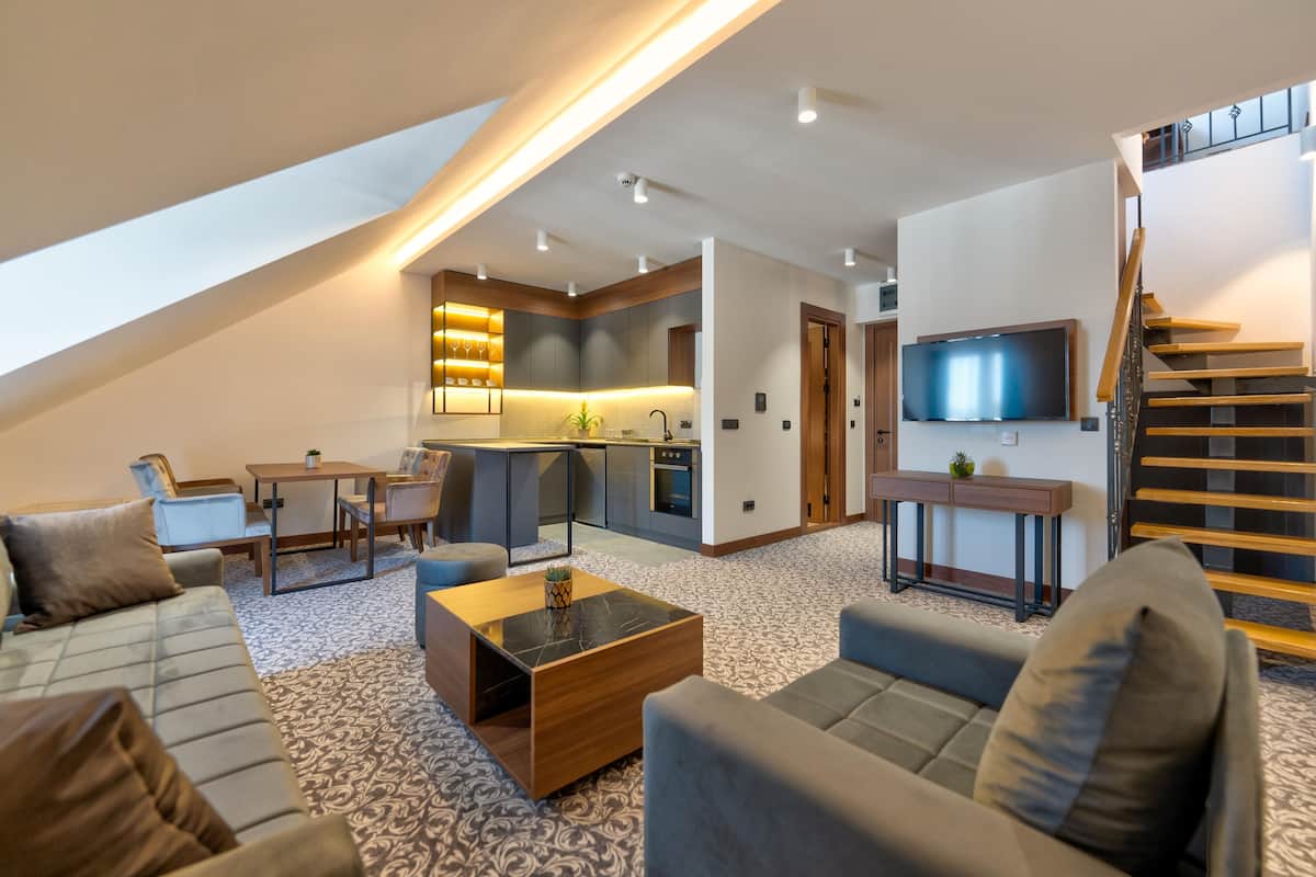 Un appartement moderne de style « loft » comprenant une cuisine, un coin repas et un salon. Il est doté d’un escalier, d’un téléviseur à écran plat et d’une grande fenêtre qui laisse entrer la lumière naturelle.