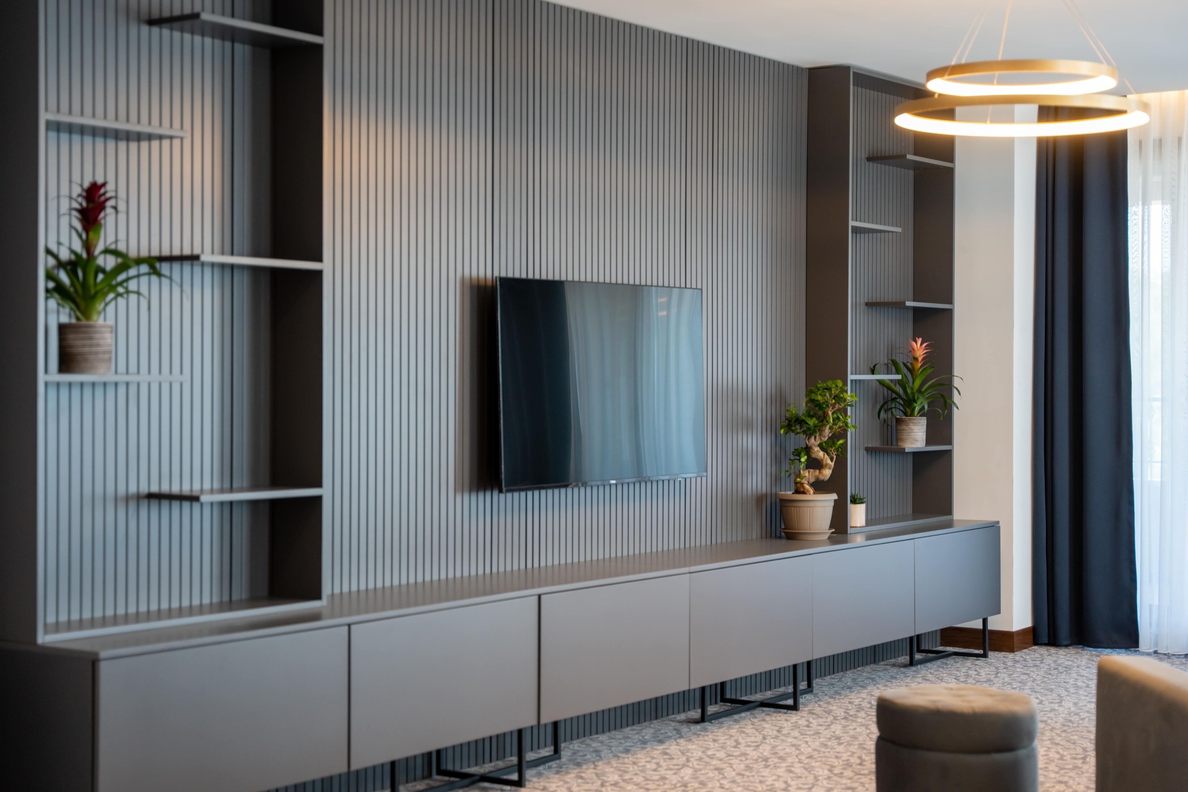 A modern living room with a flat-screen TV mounted on a wall with vertical paneling, a grey console table with cabinets, and potted plants.