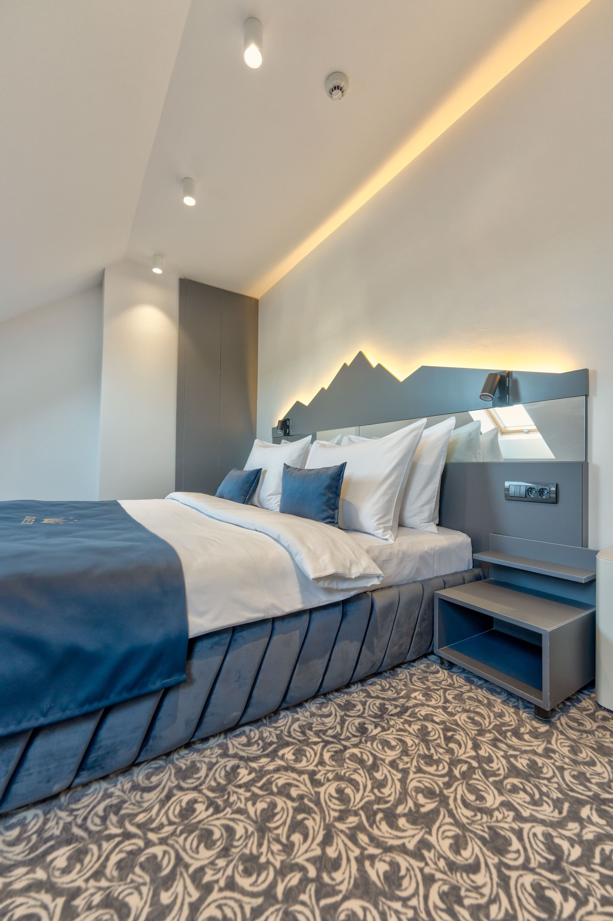Una habitación de hotel moderna con cama, mesita de noche y cabecera con diseño de montaña.