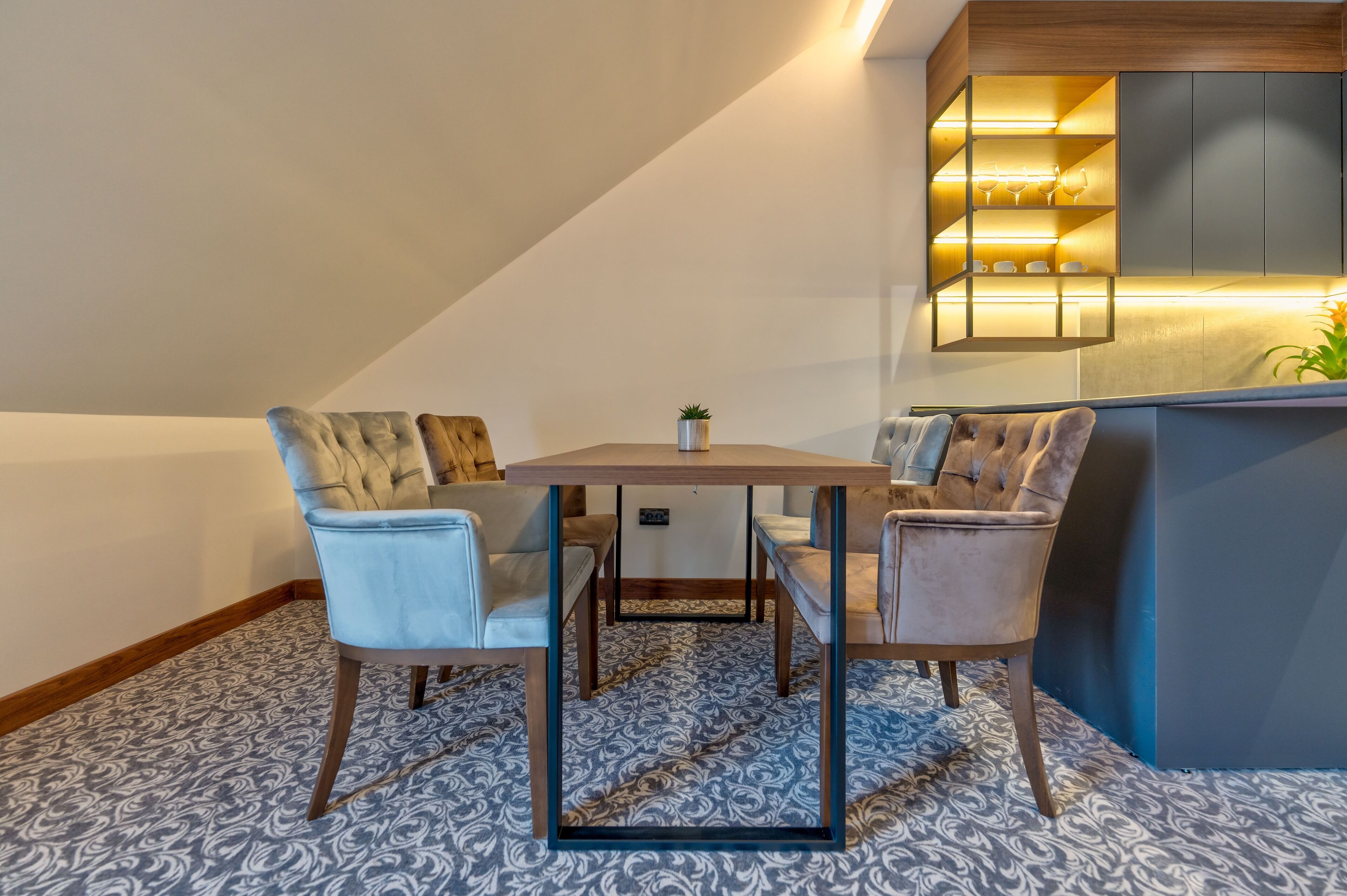 A modern loft bedroom with a dining area, a built-in kitchen with illuminated shelves, and a patterned carpet.