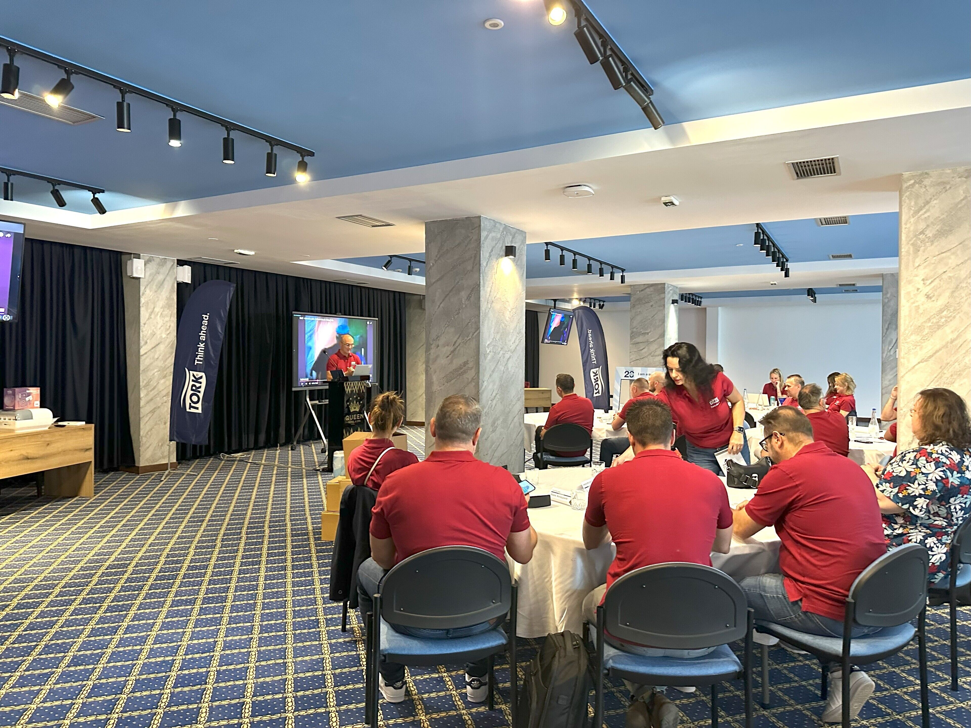 A conference room with a presentation screen, attendees seated at round tables, and a stage with a speaker.