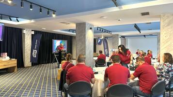 A conference room with a presentation screen, attendees seated at round tables, and a stage with a speaker.