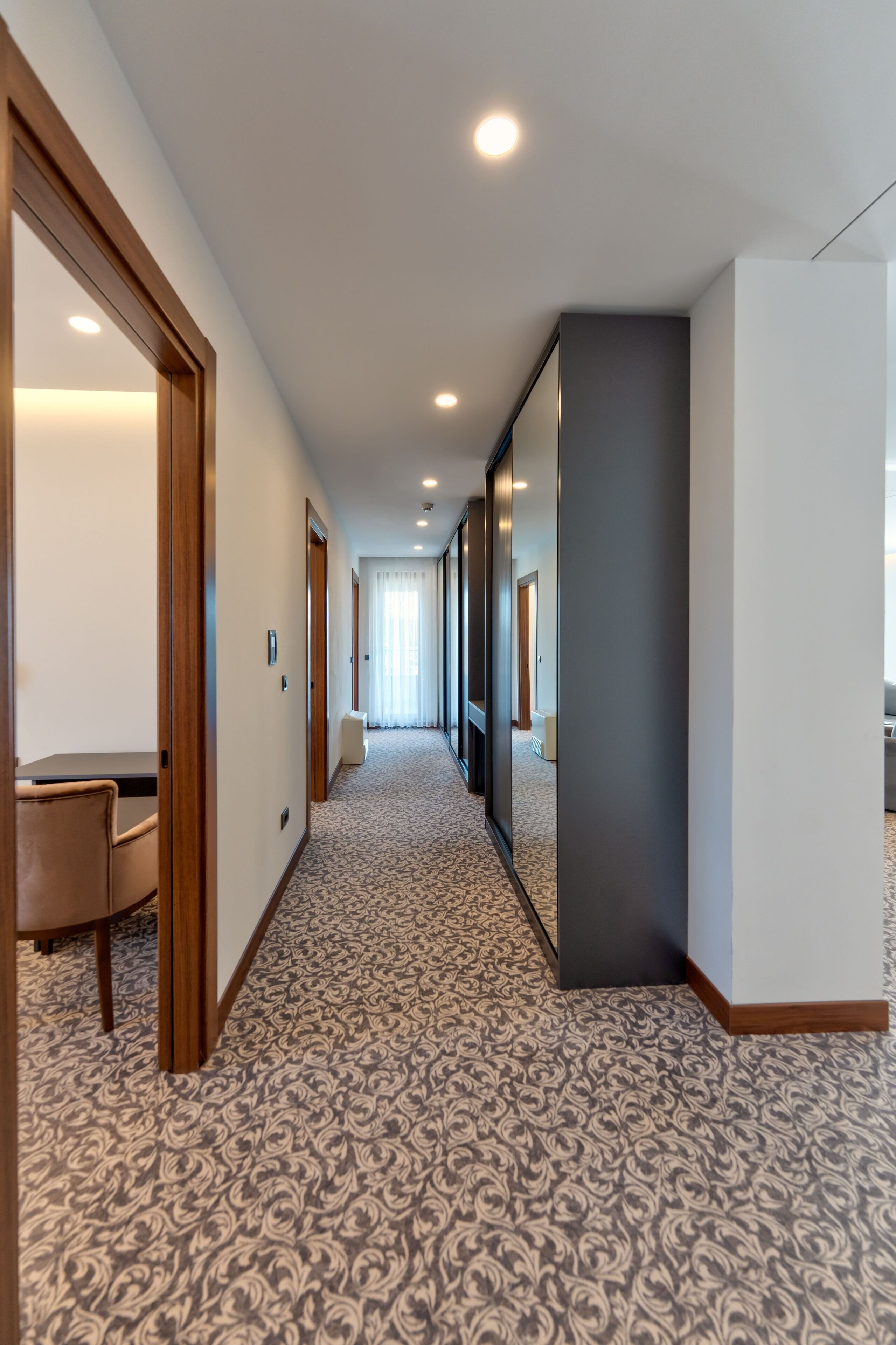Un pasillo de hotel con alfombra estampada, puertas de madera y una zona de estar con mesa y sillas.