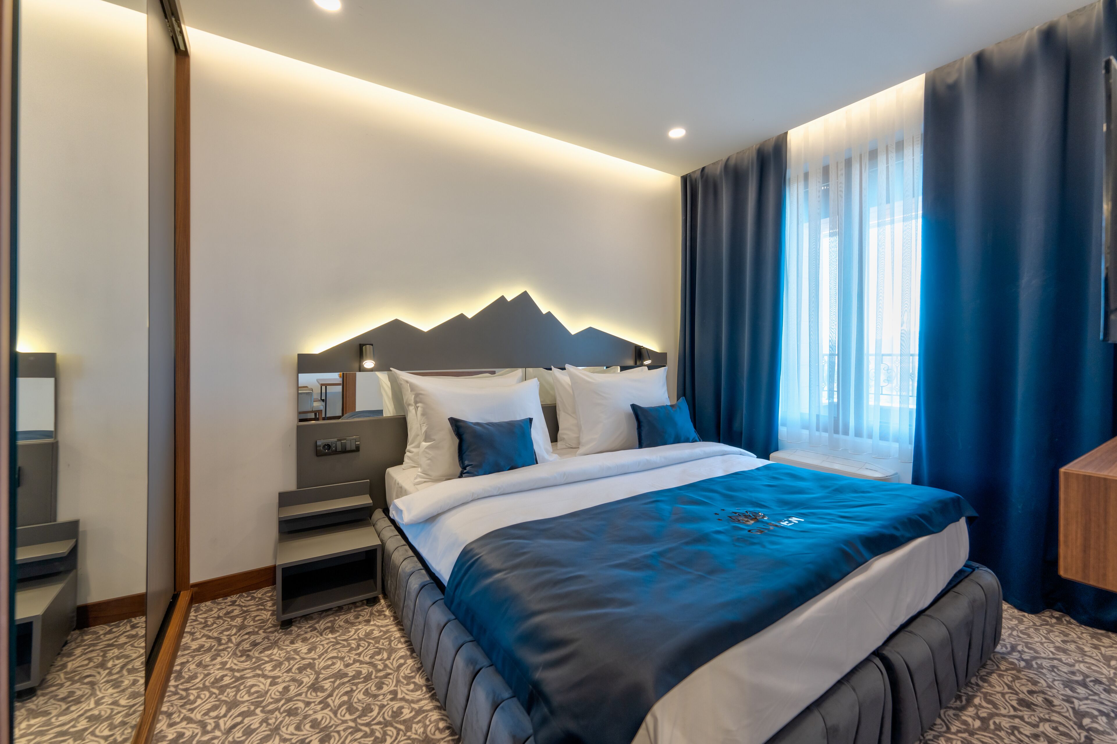 A modern hotel room with a large bed, a headboard with a mountain design, a bedside table, and a window with curtains.