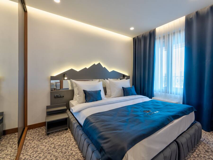 A modern hotel room with a large bed, a headboard with a mountain design, a bedside table, and a window with curtains.