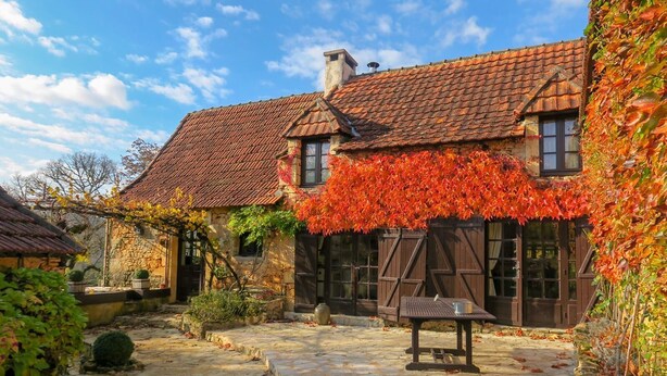 Une cour pavée de pierres, avec un banc et une table en pierre rustique, entourée de bâtiments traditionnels en pierre, dotés de volets en bois et d’un toit en tuiles.