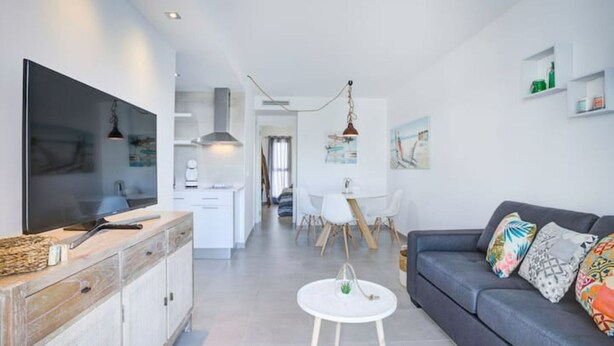 Ein modernes Wohnzimmer mit Flachbildfernseher, grauem Sofa mit bunten Kissen, weißem Couchtisch und Küchenzeile mit weißen Schränken und einem großen Fenster.
