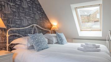 Una cama bien hecha con sábanas blancas y almohadas azules estampadas, un cabecero de metal blanco y una ventana con vista a un edificio de ladrillo.