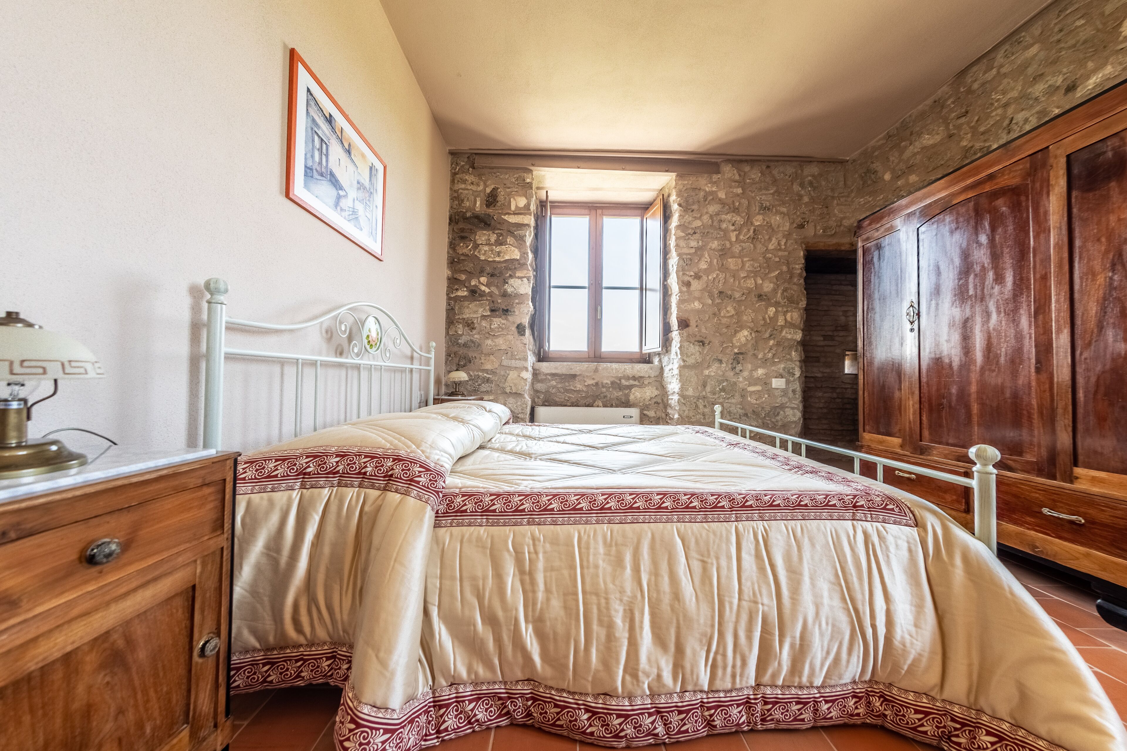 Une chambre avec un mur en pierre, une armoire en bois, un lit recouvert d’une couvre-lit à motifs et une fenêtre donnant sur un paysage agréable.