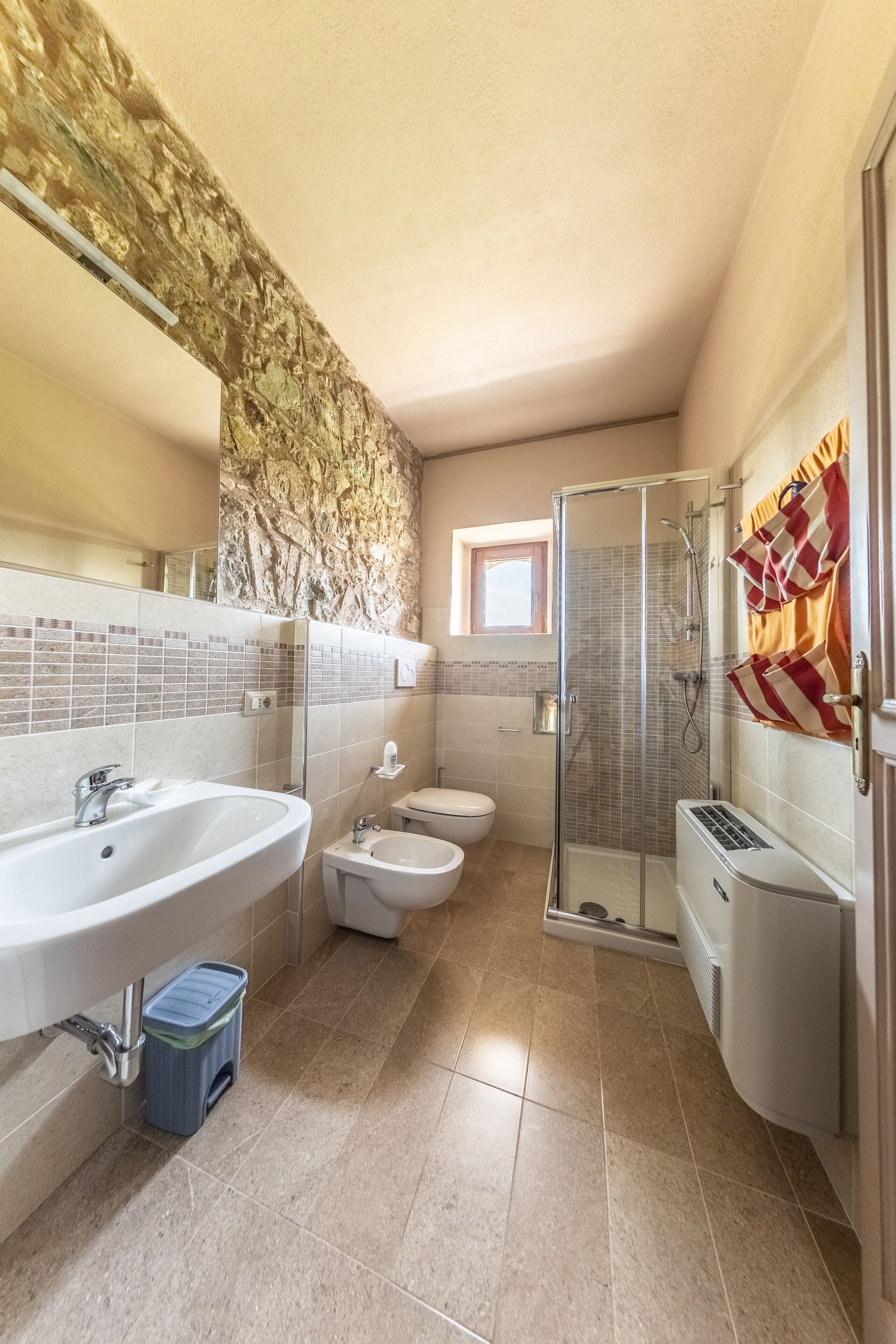 Un baño con pared de piedra, un lavamanos grande, un bidé, una cabina de ducha con mampara de vidrio y un inodoro.