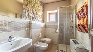 Un baño con pared de piedra, un lavamanos grande, un bidé, una cabina de ducha con mampara de vidrio y un inodoro.