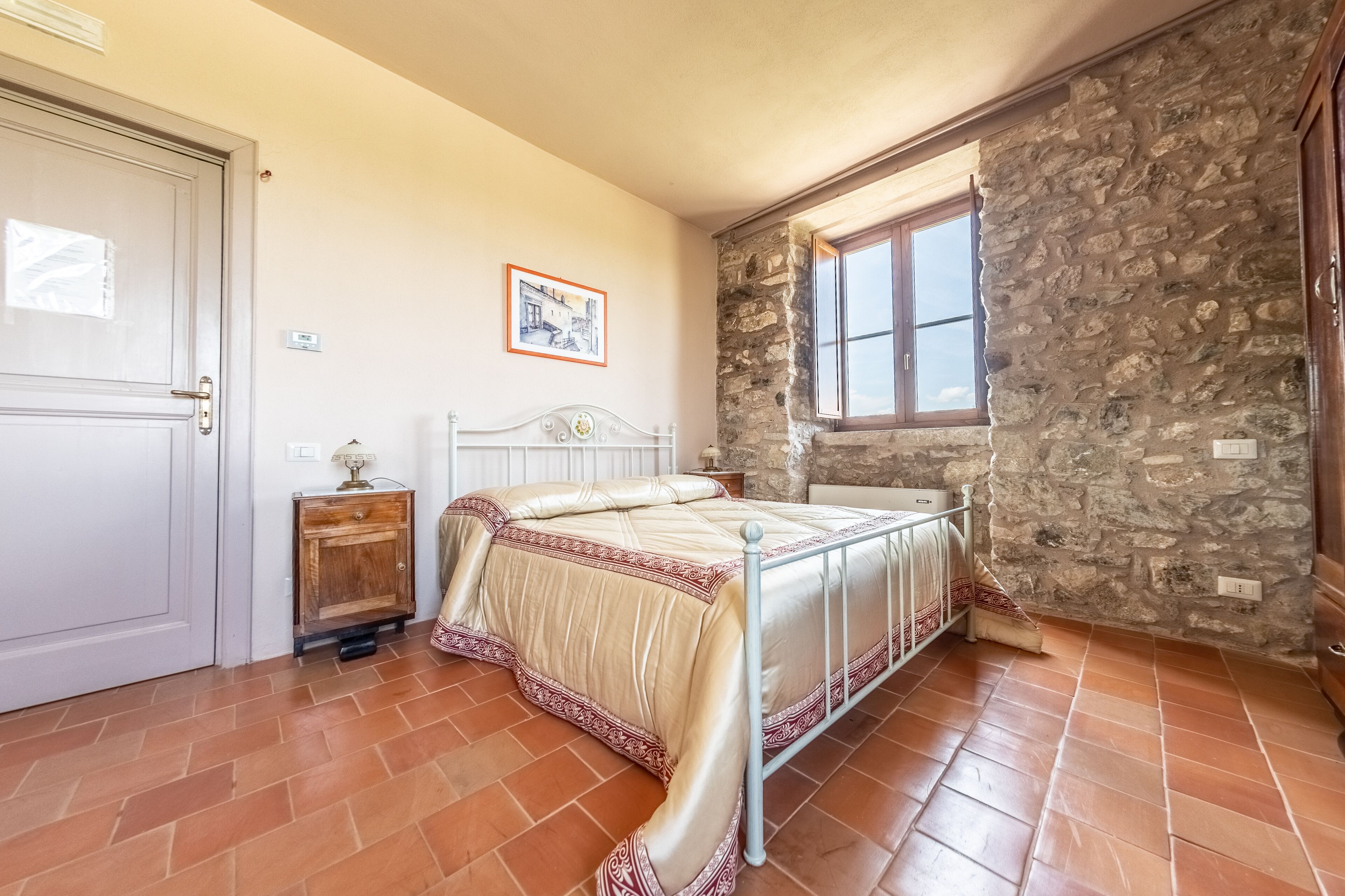 Ein Schlafzimmer mit Steinwand, einem Bett mit gemustertem Überwurf, einem Nachttisch aus Holz und einem Fenster mit Blick ins Freie.