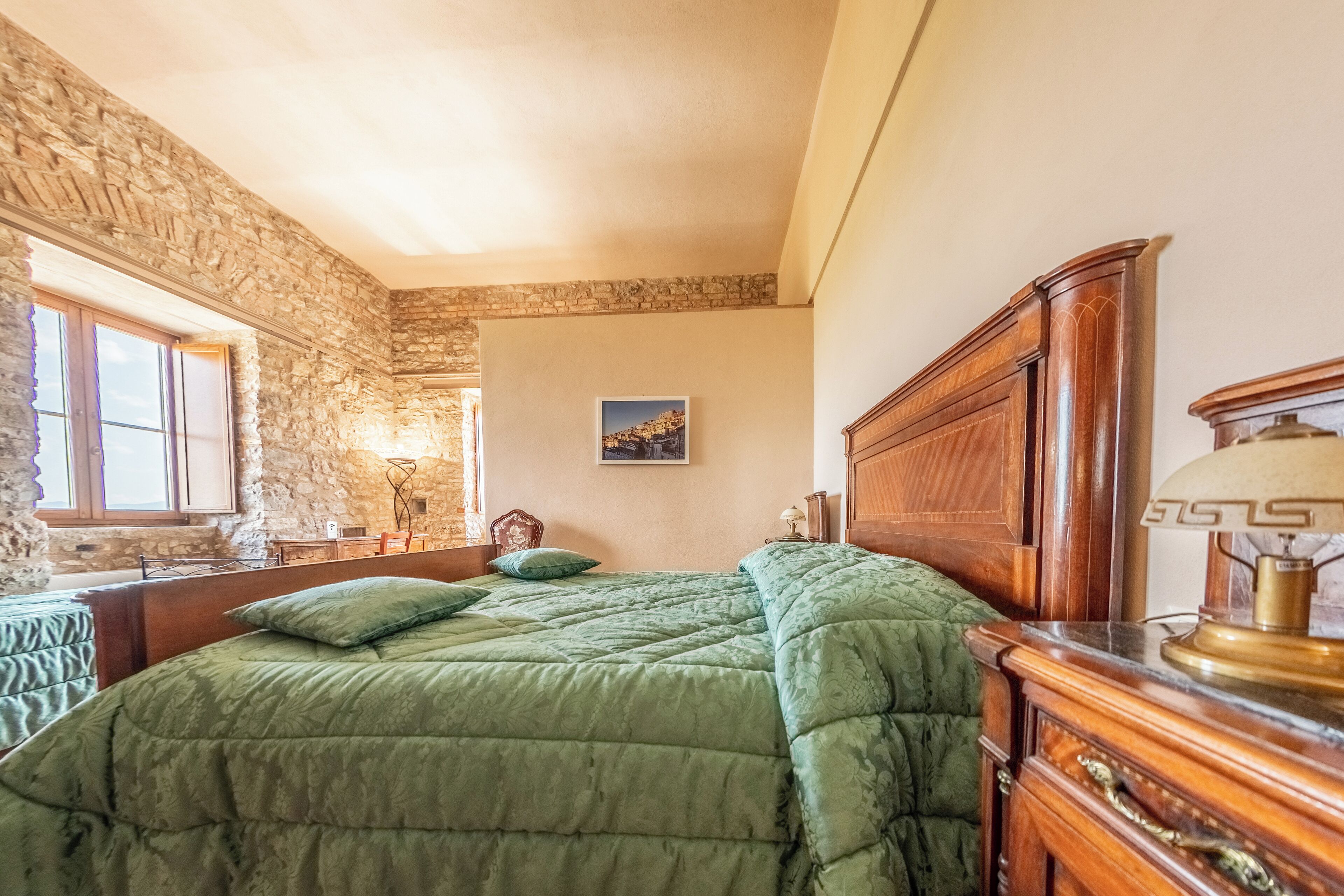 Un dormitorio con pared de piedra, cama de madera, colcha verde, mesita de noche, lámpara y un cuadro en la pared.