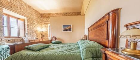 Un dormitorio con pared de piedra, cama de madera, colcha verde, mesita de noche, lámpara y un cuadro en la pared.