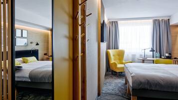 A hotel room with two beds, a yellow armchair, a desk with a lamp, and a large window with curtains.