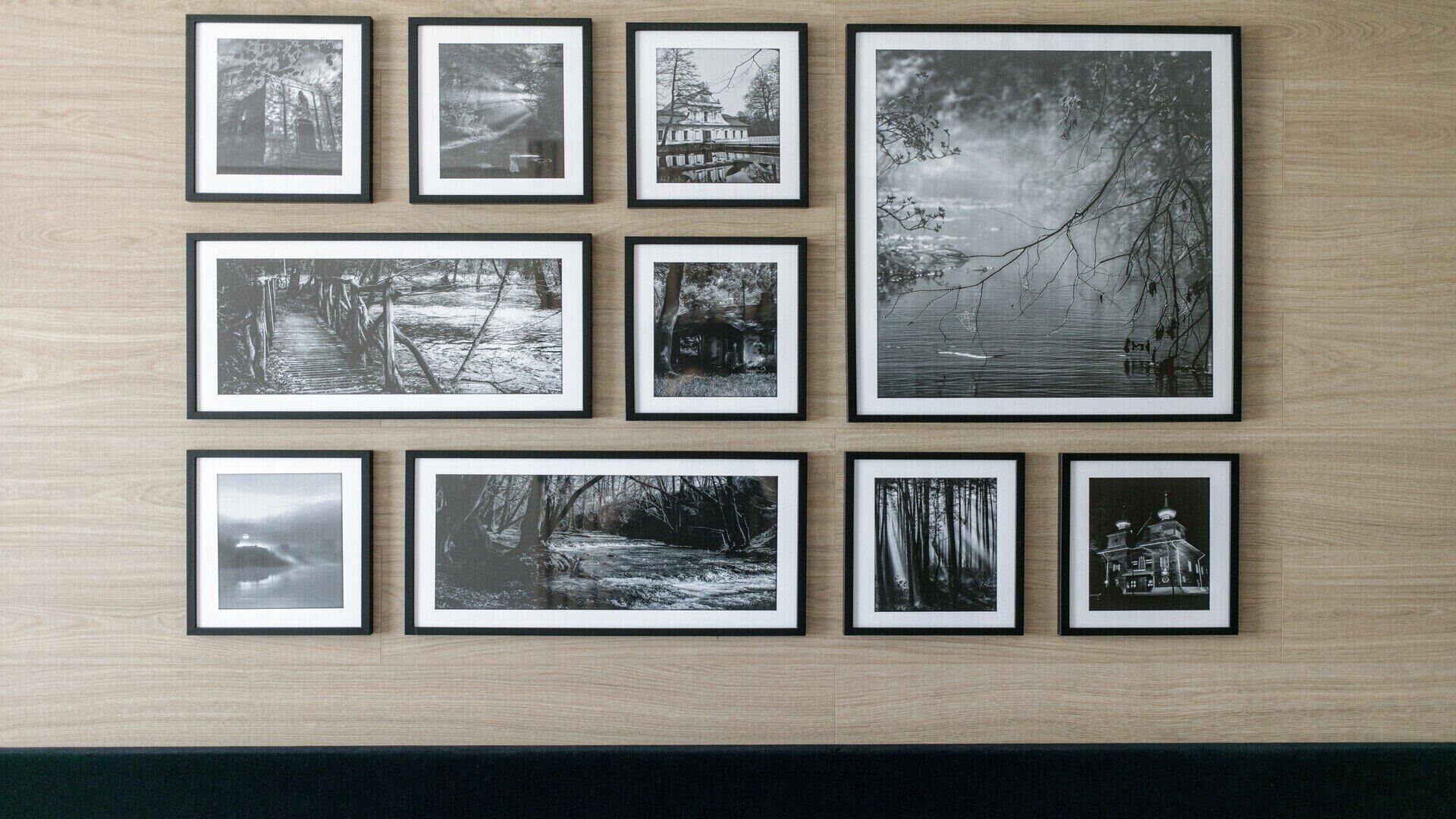 A wall with nine framed black and white photographs of various outdoor scenes, including trees, a bridge, and a house by a body of water.