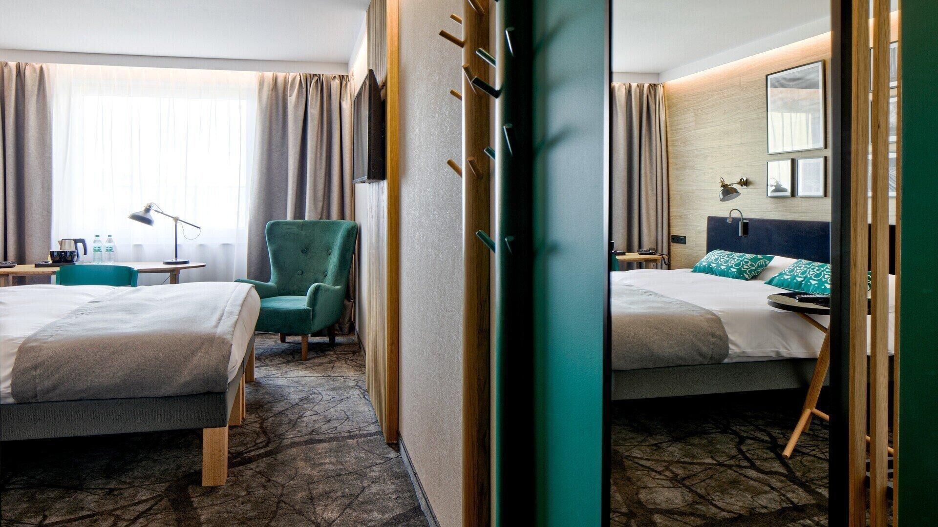 A hotel room with two beds, a green armchair, a desk with a lamp, and a large mirror.