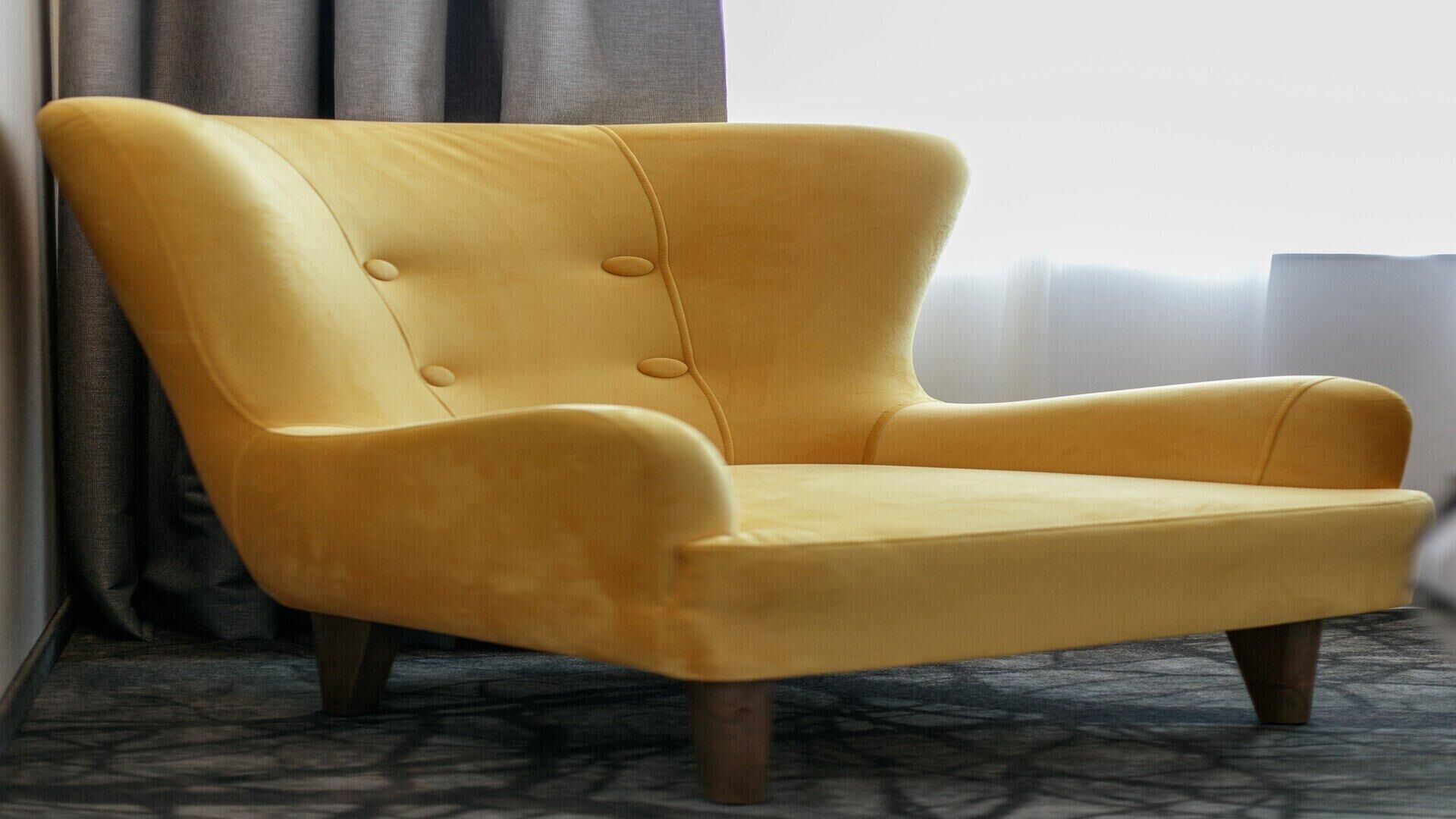 A yellow armchair with a high back and button details, positioned in a room with a patterned carpet and curtains.