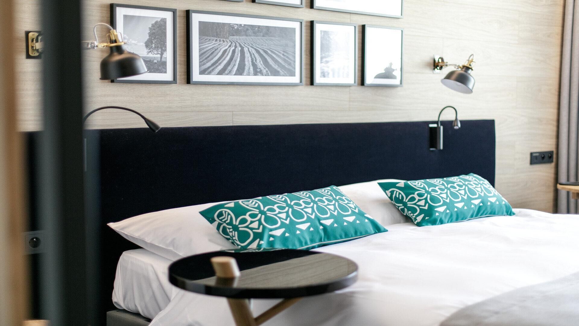 Habitación de hotel con una cama, dos almohadas, una mesita de noche y una pared decorada con cuadros.