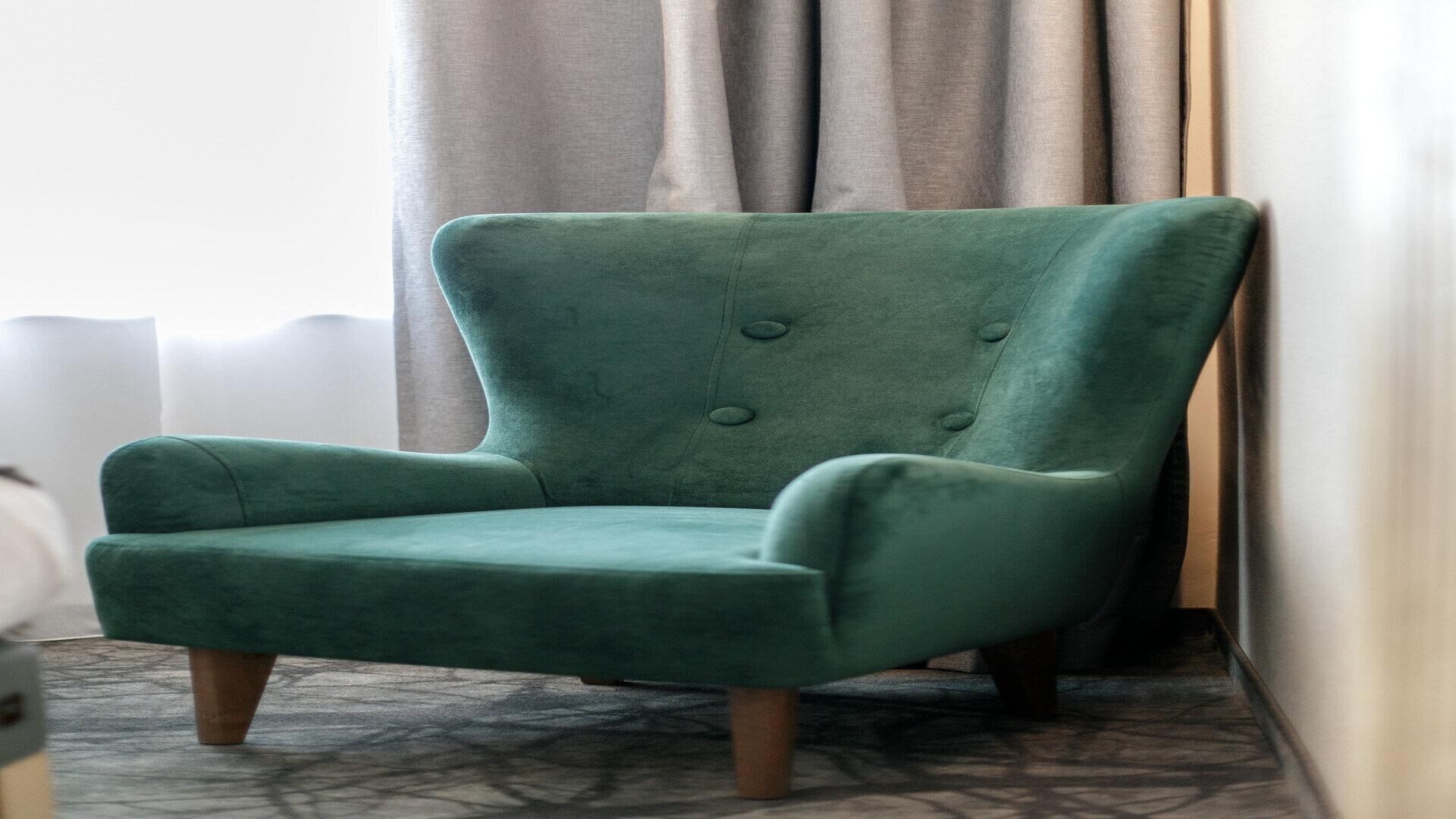 Un sillón verde con patas de madera, ubicado frente a una pared con ventana y cortinas.