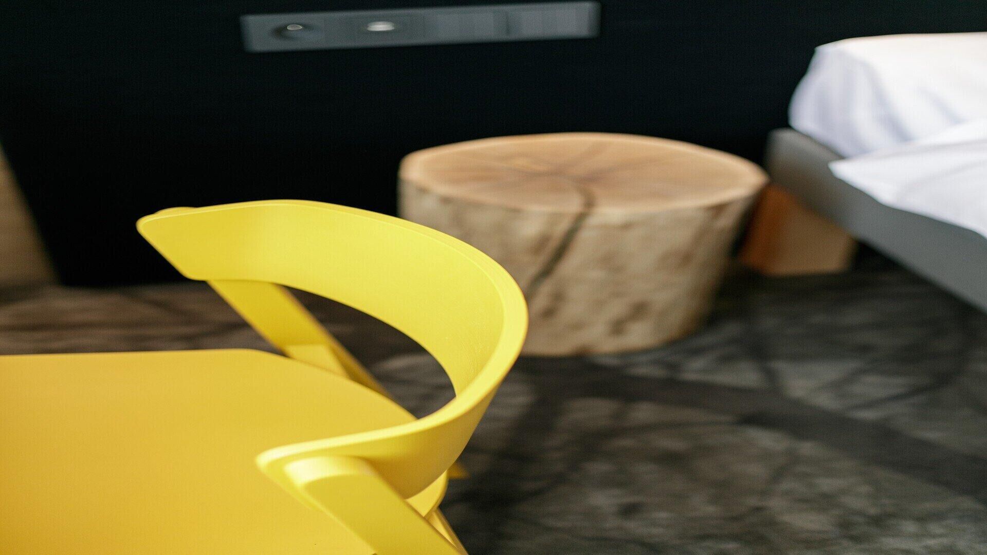 Une chaise jaune avec un dossier incurvé, dans une pièce dont le sol est foncé, avec un tabouret en bois et un lit recouvert d’une couverture blanche.