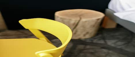 A yellow chair with a curved backrest in a room with a dark floor, a wooden stool, and a bed with a white cover.