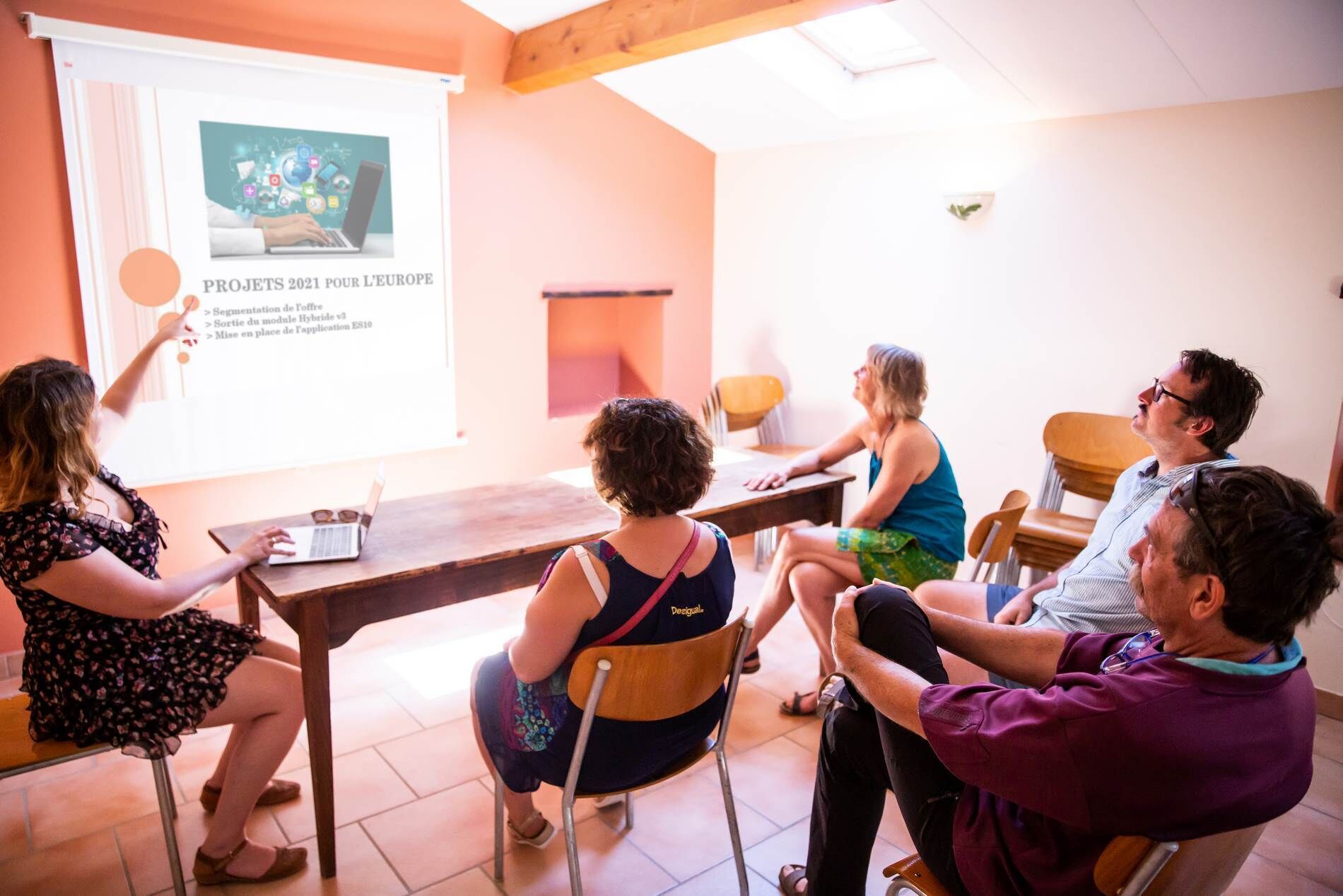 Um grupo de pessoas em uma sala de reuniões, com tela de apresentação, mesas e cadeiras de madeira.