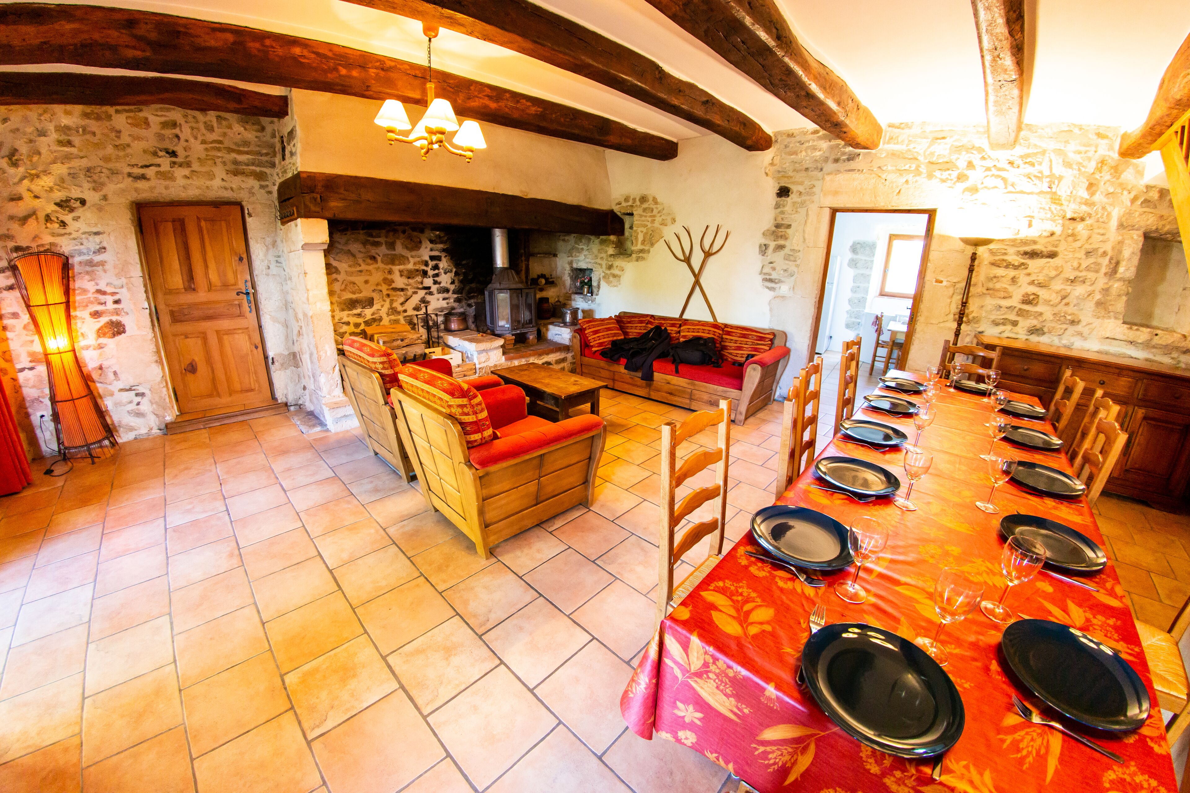 Un comedor rústico con una larga mesa lista para una comida, paredes de piedra, vigas de madera y una zona de estar acogedora.
