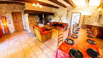 Un comedor rústico con una larga mesa lista para una comida, paredes de piedra, vigas de madera y una zona de estar acogedora.